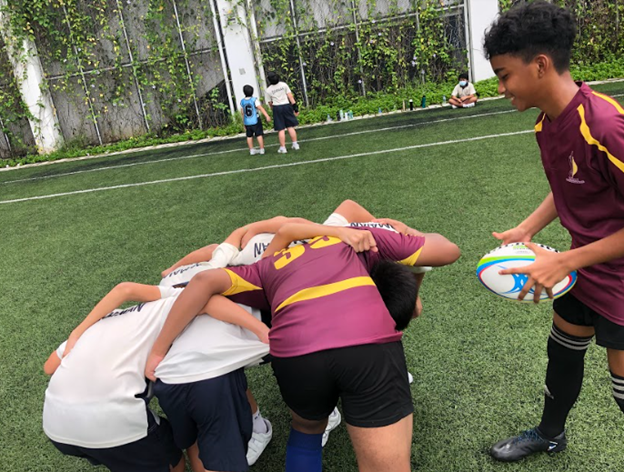 Young players in maroon jerseys practice a rugby scrum on a green turf field while one boy holds a rugby ball.